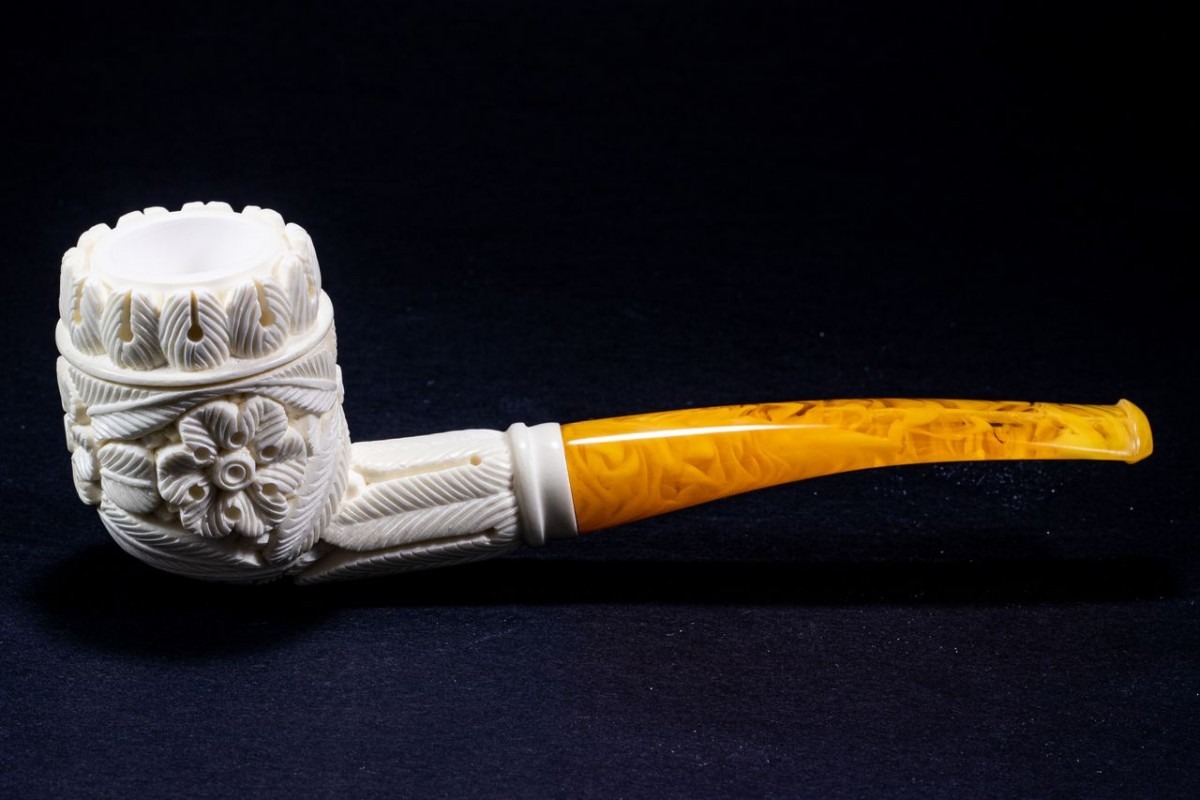 Intricately carved Meerschaum Quarter Bent Billiard pipe with floral and leaf motifs on a white bowl and a gently curved amber stem, set against a dark background.